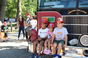 Photo of kids wearing fire helmets sitting on the front bumper of a fire truck